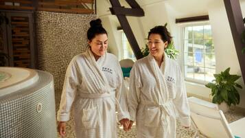Two individuals in white robes with "DEEP NATURE" text, walking in a spa-like setting with a stone wall, a large bathtub, and a wooden ceiling structure.