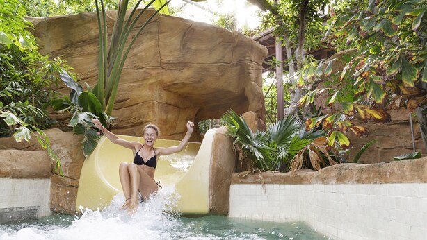 A person sliding down a water slide in a pool area surrounded by rocks and tropical plants. - Center Parcs Le Lac d'Ailette (Chamouille)