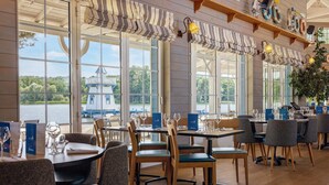 A restaurant with large windows, wooden furniture, and a view of a lake and a house. - Center Parcs Le Lac d'Ailette (Chamouille)