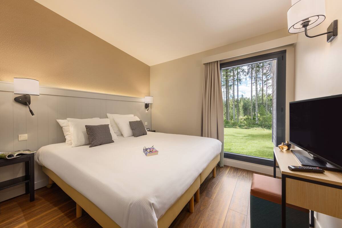 A hotel room with a large bed, a desk with a TV, and a window with a view of trees.