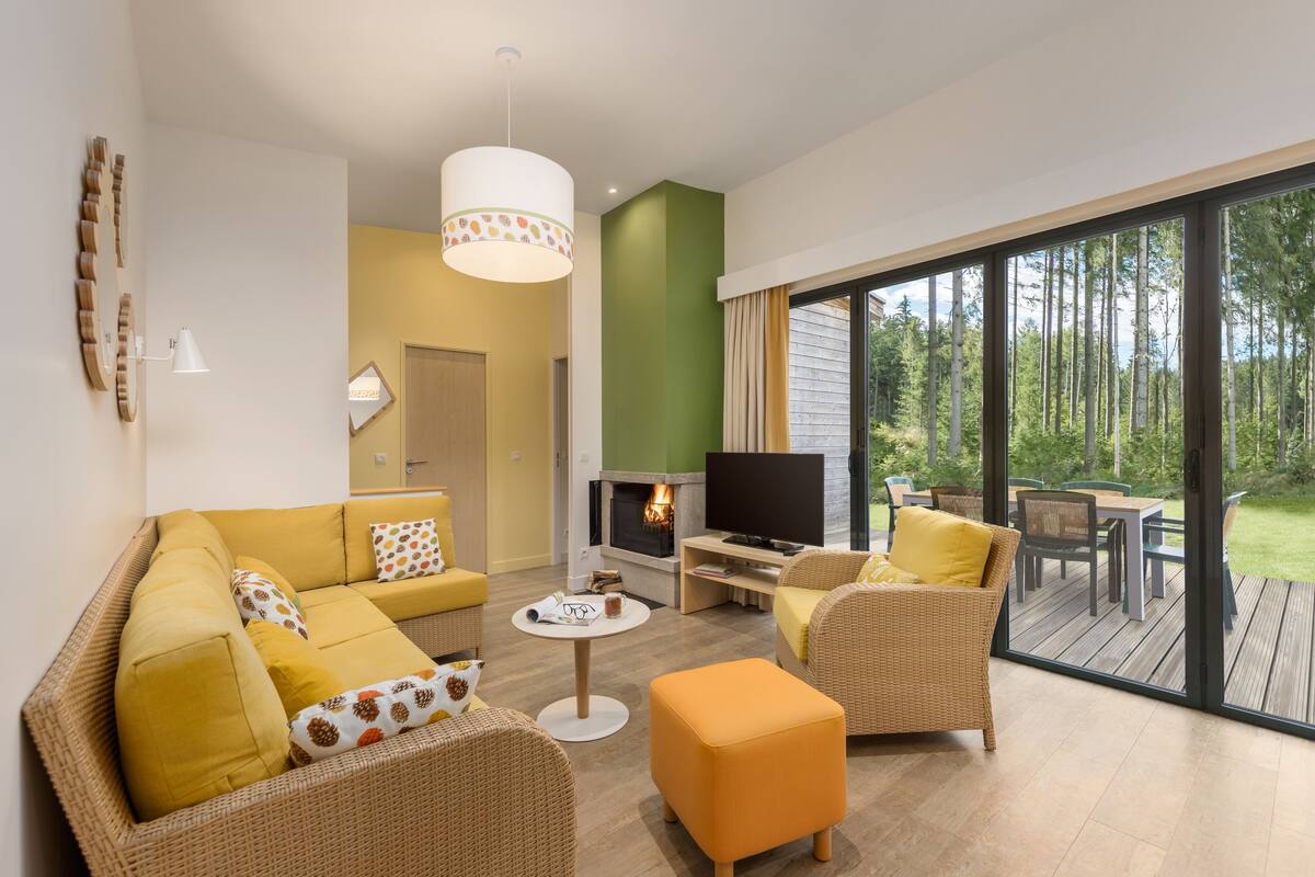 A modern living room with a sofa, ottoman, and coffee table, a fireplace, and a large window overlooking a deck.