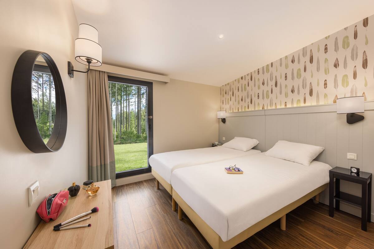 A hotel room with two beds, a large mirror, a desk with makeup and a handbag, and a view of greenery outside.