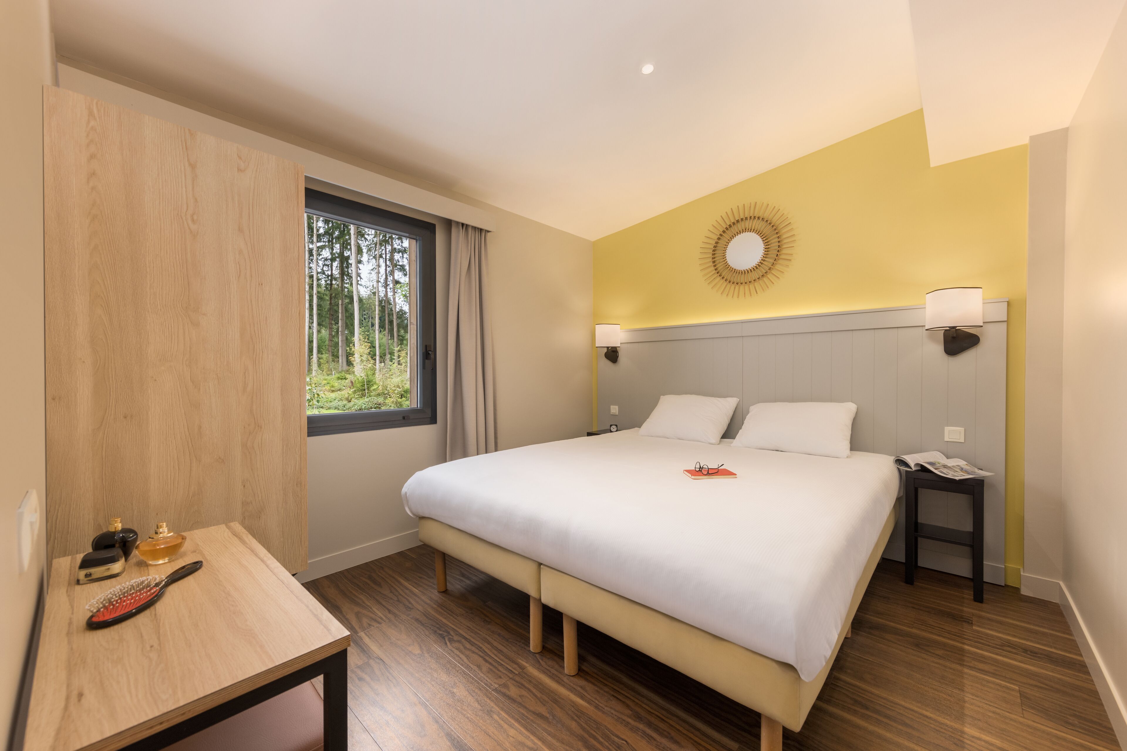 Moderni hotellihuone, jossa on suuri sänky, yöpöytälamput, puinen yöpöytä ja näkymä puita kohti ikkunasta.