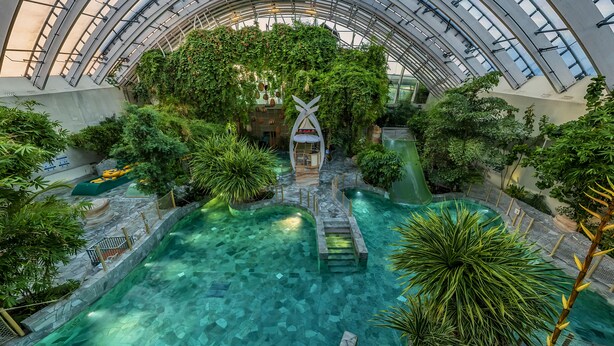 An indoor atrium with a glass ceiling, a central water feature, and lush greenery. - Center Parcs Les Trois Forêts (Fraquelfing)