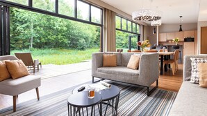 A modern living room with a grey sofa, a coffee table, and a dining area with a table and chairs. Large windows provide a view of a forested area. - Center Parcs Les Trois Forêts (Fraquelfing)