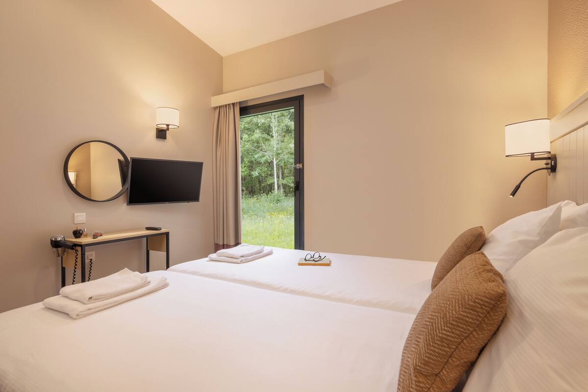 A hotel room with two beds, a TV, a desk with a lamp, and a window with a view of greenery.