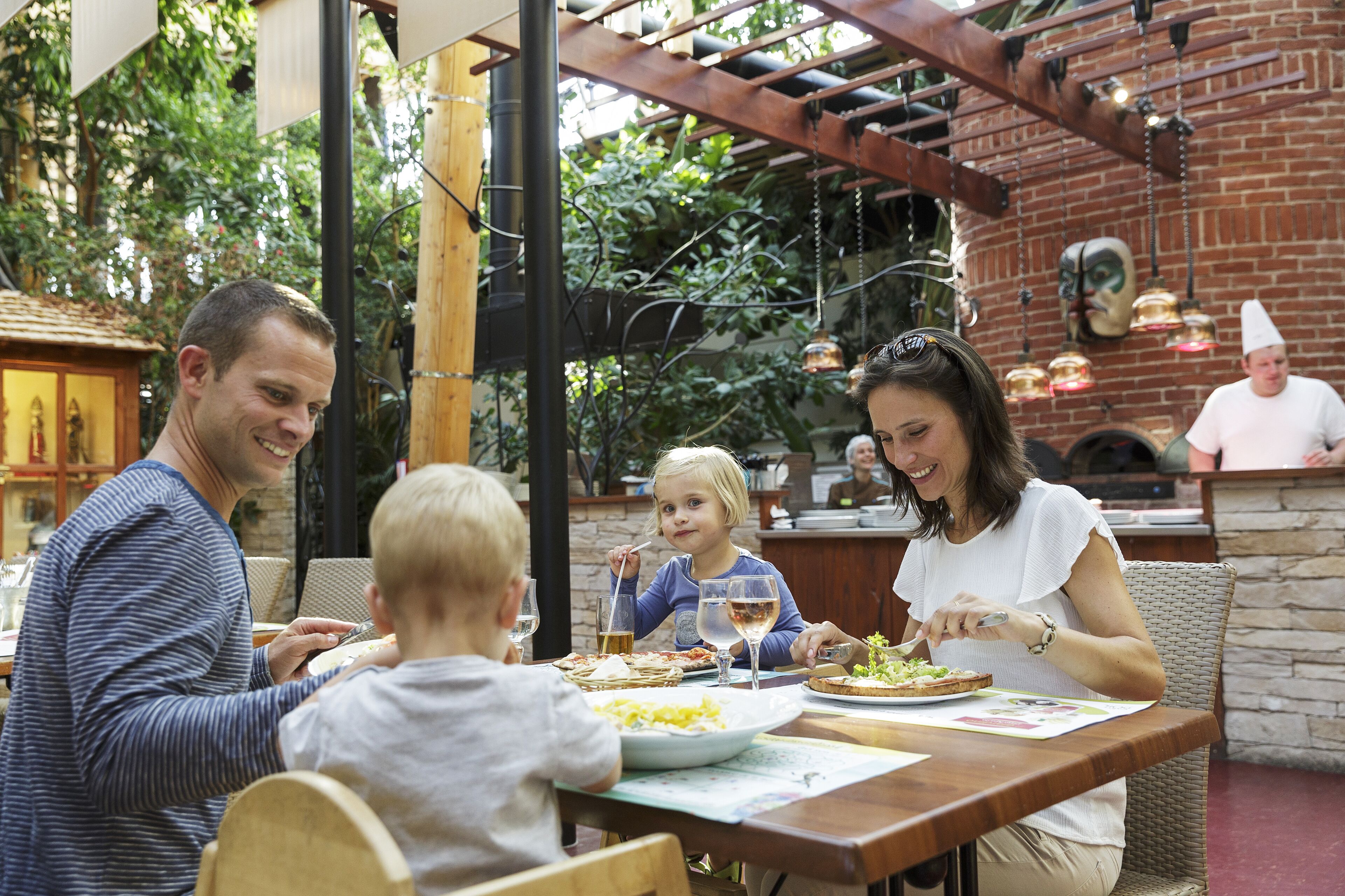 A family dining at an outdoor restaurant with a brick wall and greenery in the background.