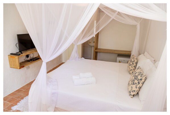 A neatly made bed with white bedding and a canopy, a bedside table with a remote control, and a small shelf with a television.