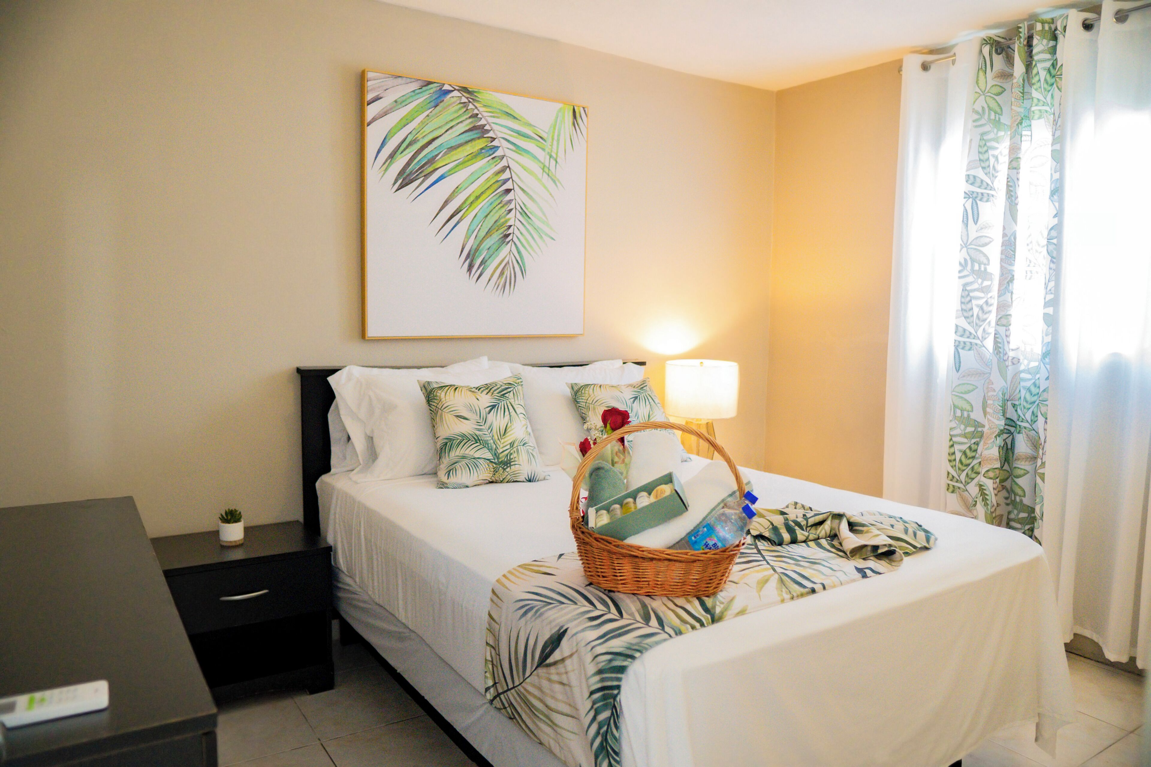 A hotel room with a bed, a nightstand, a lamp, a wall art of a palm leaf, and a window with curtains.