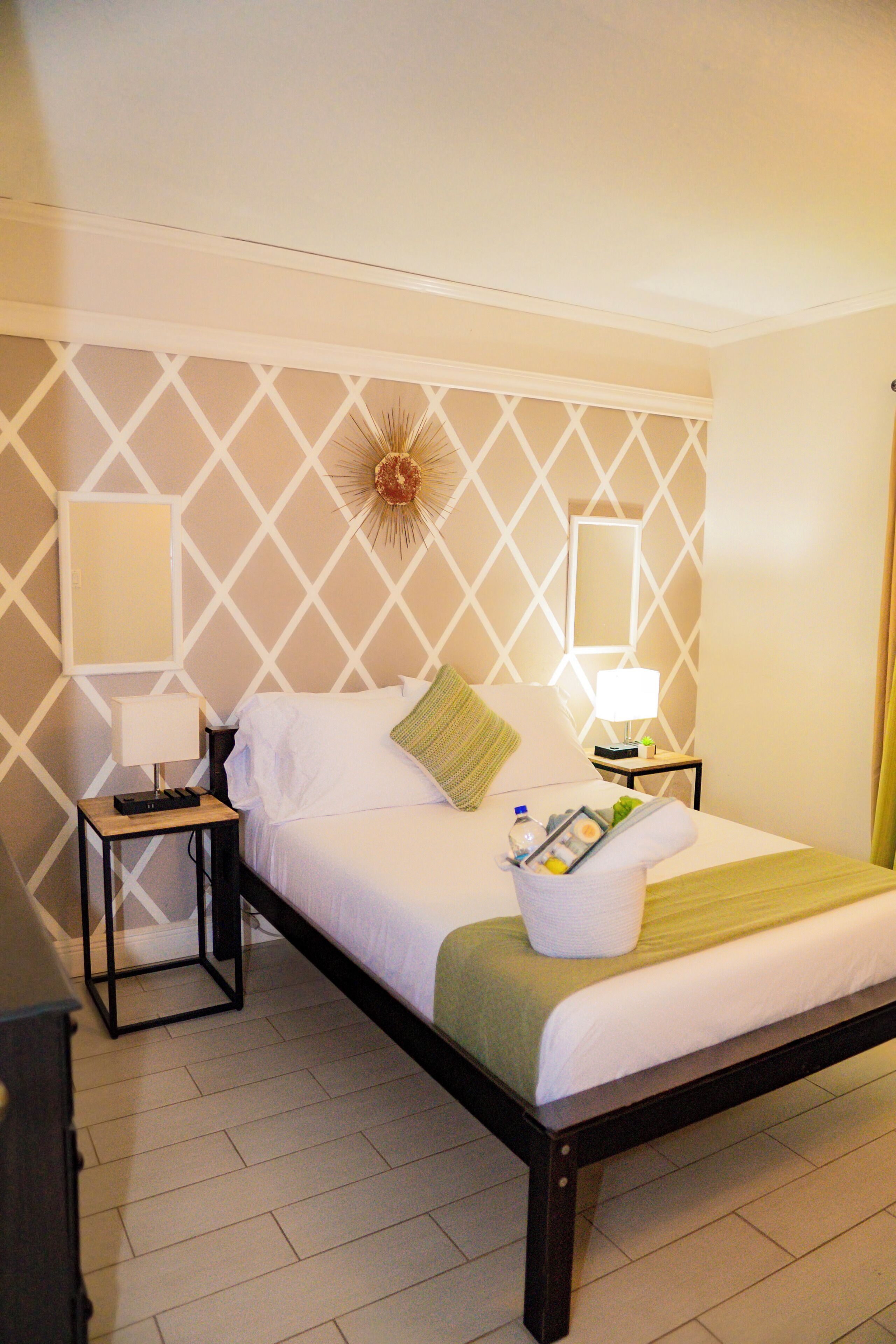 Cama bem arrumada com lençóis brancos, um cobertor verde e uma mesa de cabeceira com abajur e um pequeno bandeja de produtos de higiene pessoal.