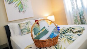 Une chambre d’hôtel avec un lit, un panier en osier contenant des articles de toilette, un tableau représentant une feuille de palmier et une lampe.