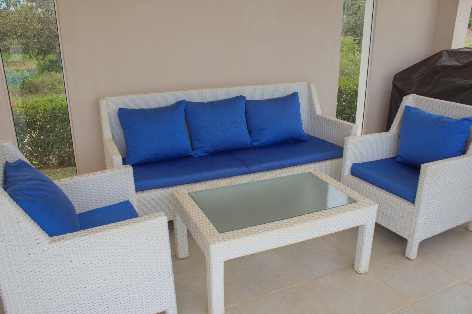 A white wicker sofa set with blue cushions, a glass-top coffee table, and a view of greenery through large windows.