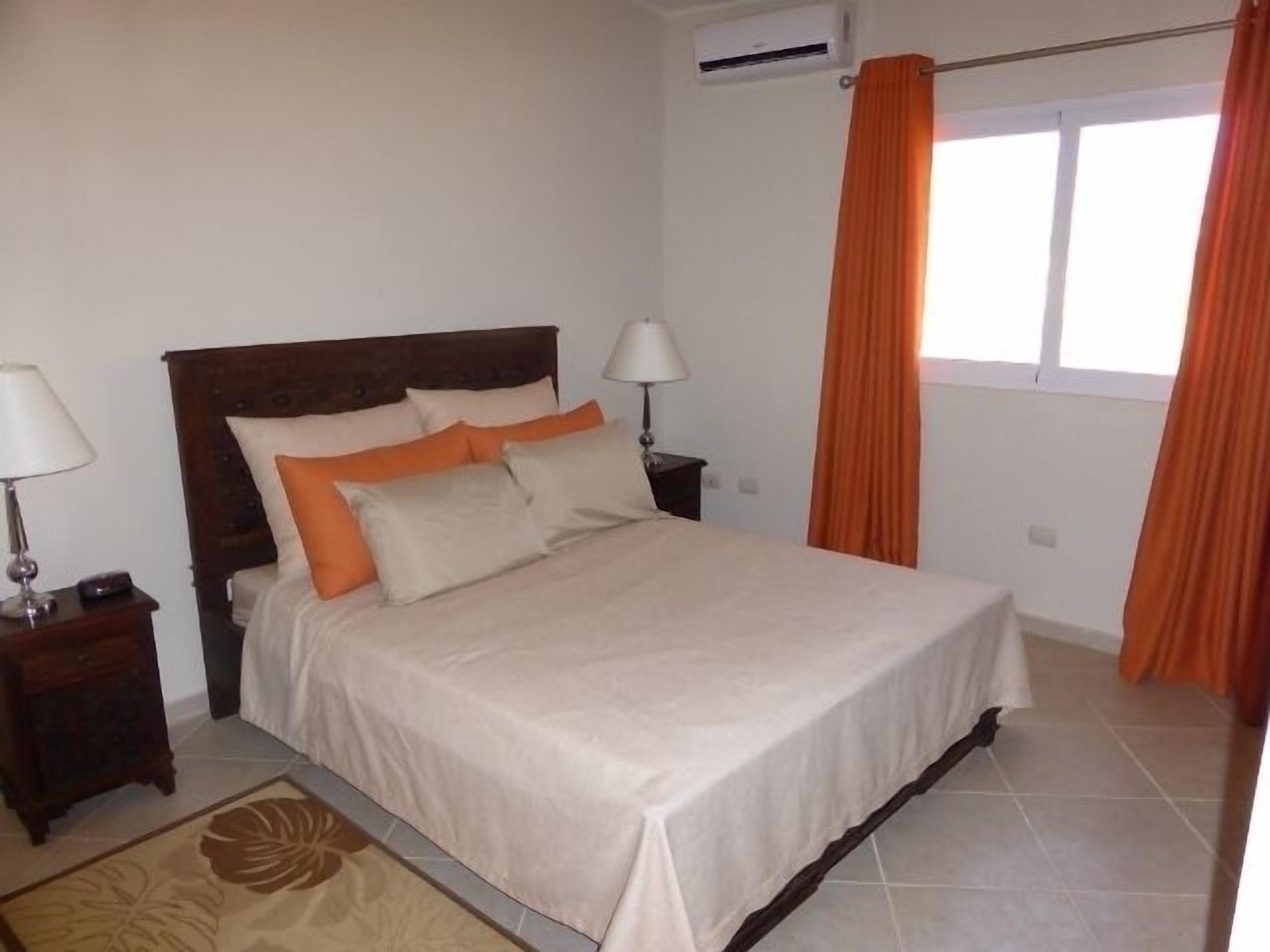 Una cama bien hecha con cabecera de madera, dos lámparas de noche y una ventana con cortinas naranjas.