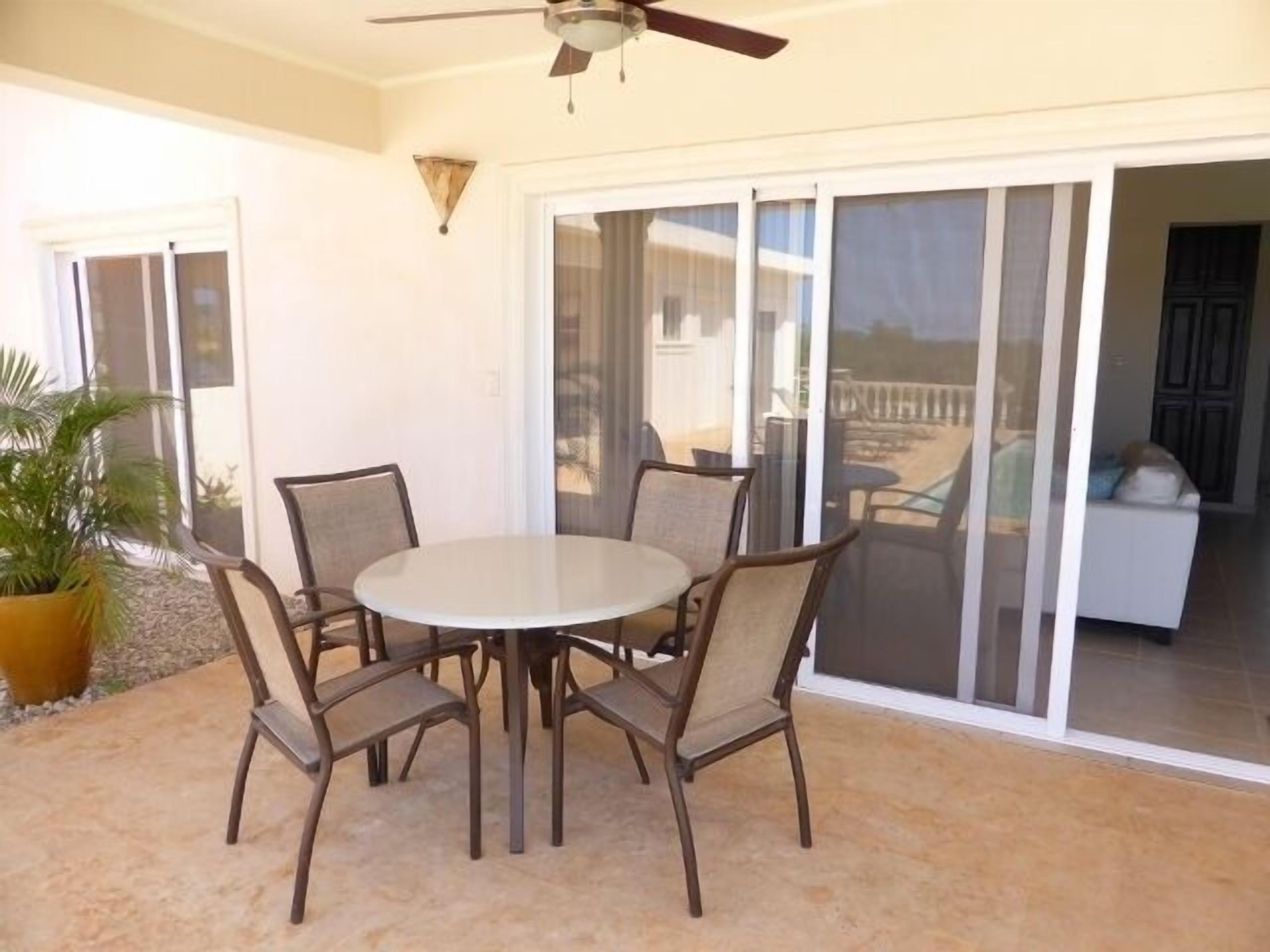 Une terrasse avec une table ronde et quatre chaises, un ventilateur de plafond et une porte-fenêtre coulissante donnant sur un espace extérieur.
