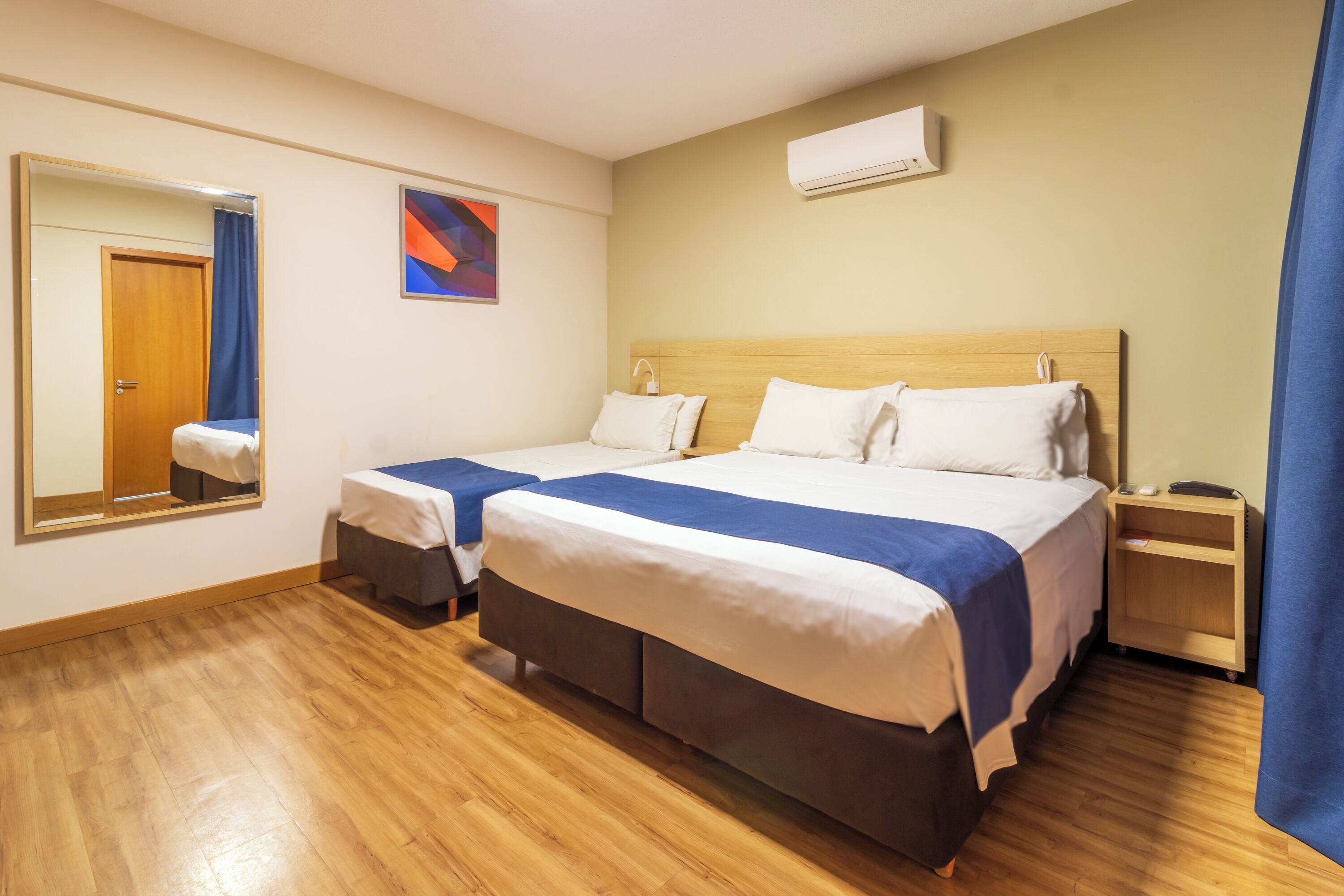 A hotel room with two beds, a wooden floor, a large mirror, a wall-mounted air conditioner, and a small bedside table.