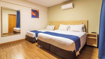 A hotel room with two beds, a wooden floor, a large mirror, a wall-mounted air conditioner, and a small bedside table.