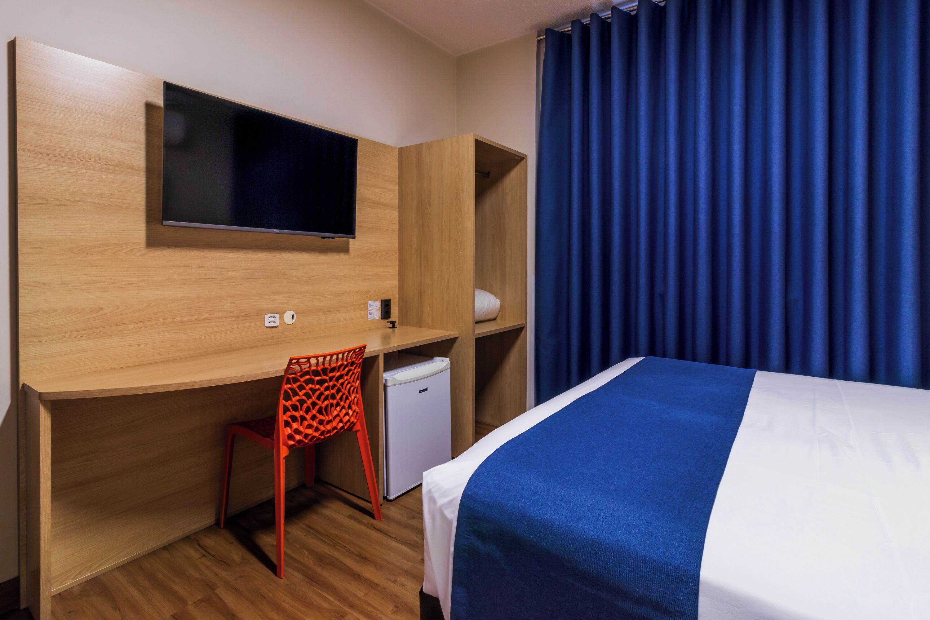 A hotel room with a bed, a desk with a chair, a television, and a small refrigerator.