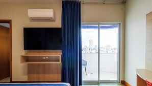Una habitación de hotel moderna con una cama, un televisor de pantalla plana, un estante de madera, una puerta de cristal corredera a un balcón y un equipo de aire acondicionado.