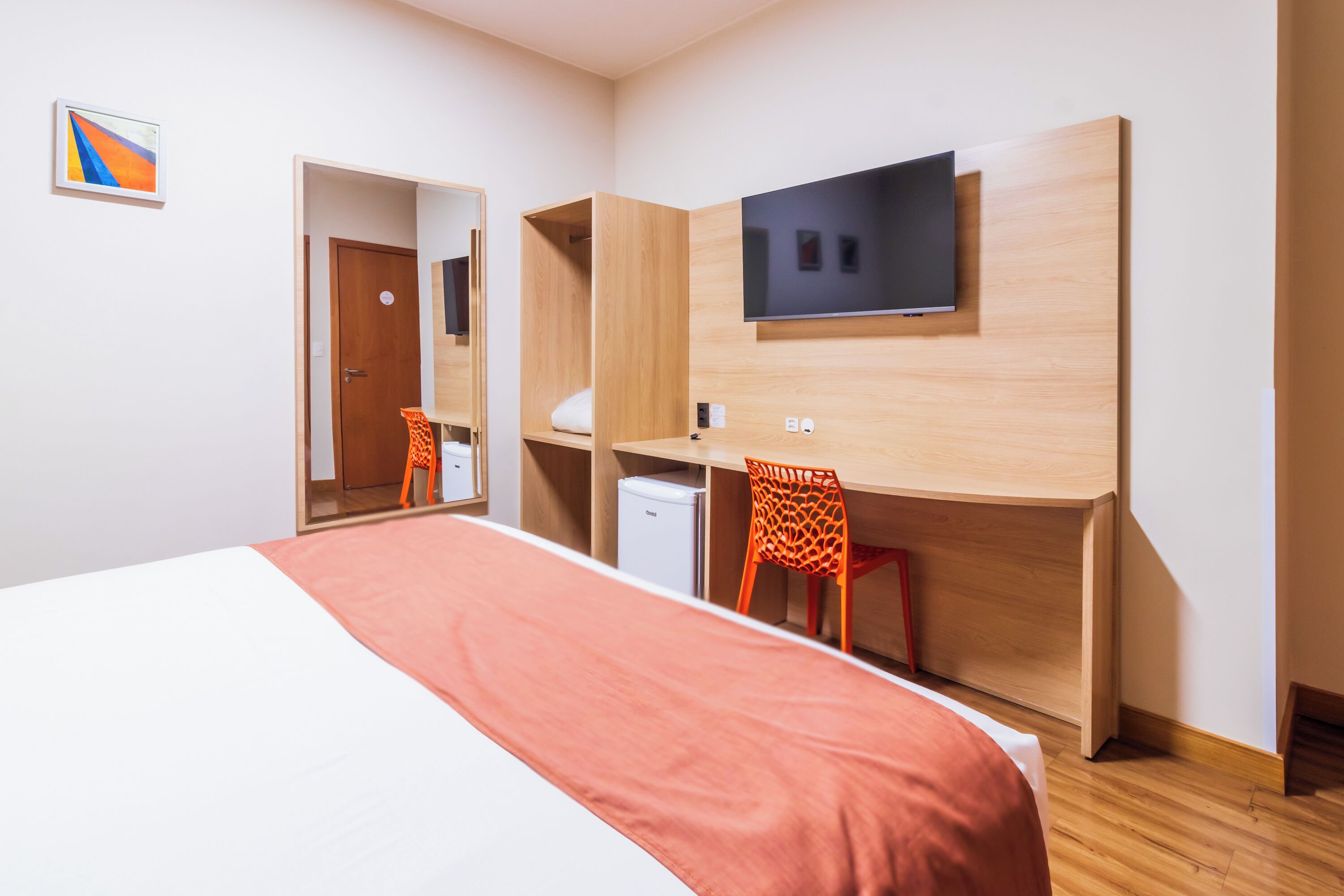 A hotel room with a bed, a desk with a chair, a television, and a small refrigerator.