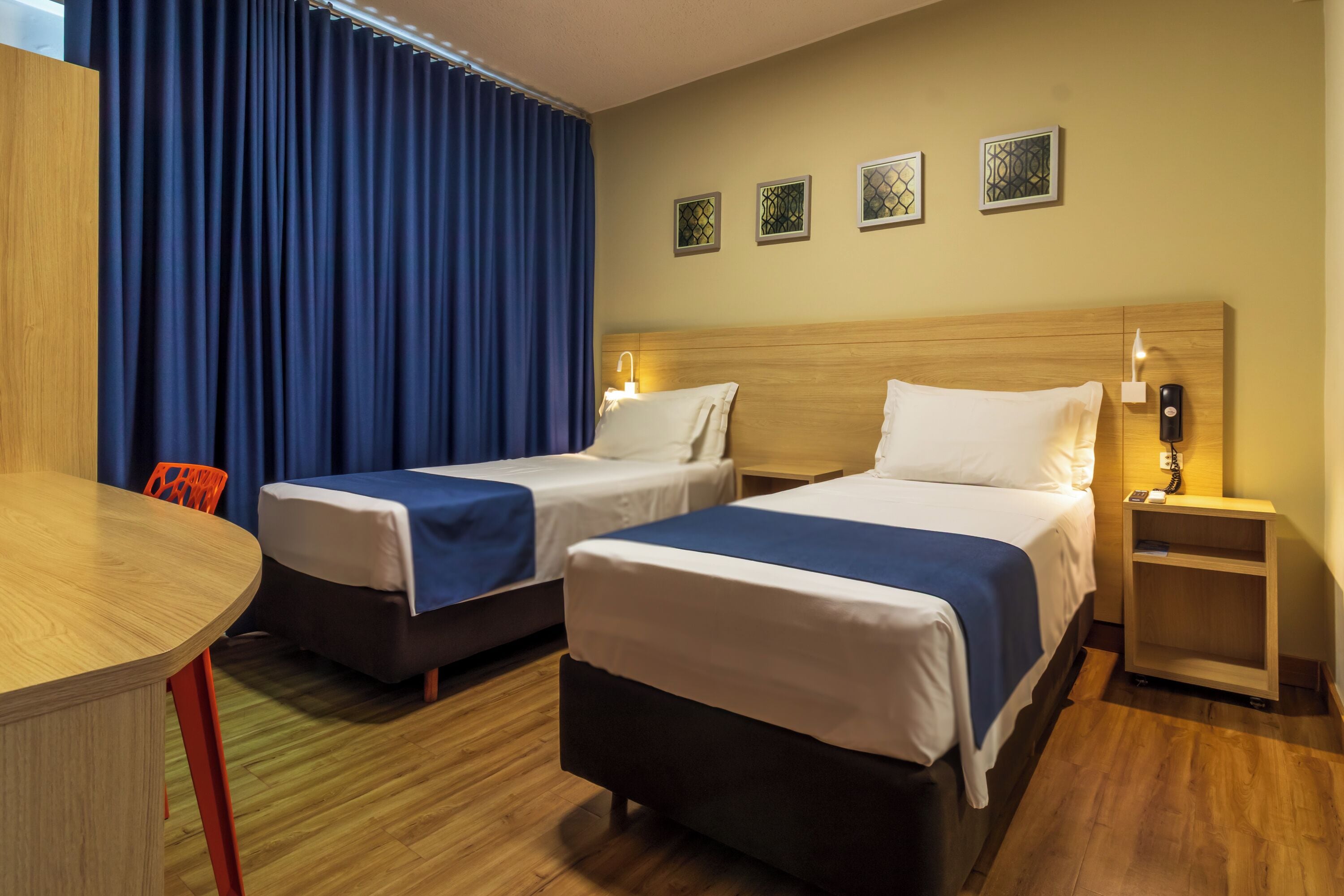 A hotel room with two beds, a wooden headboard, a small table, and a chair. There are four wall-mounted light fixtures and a window with blue curtains.