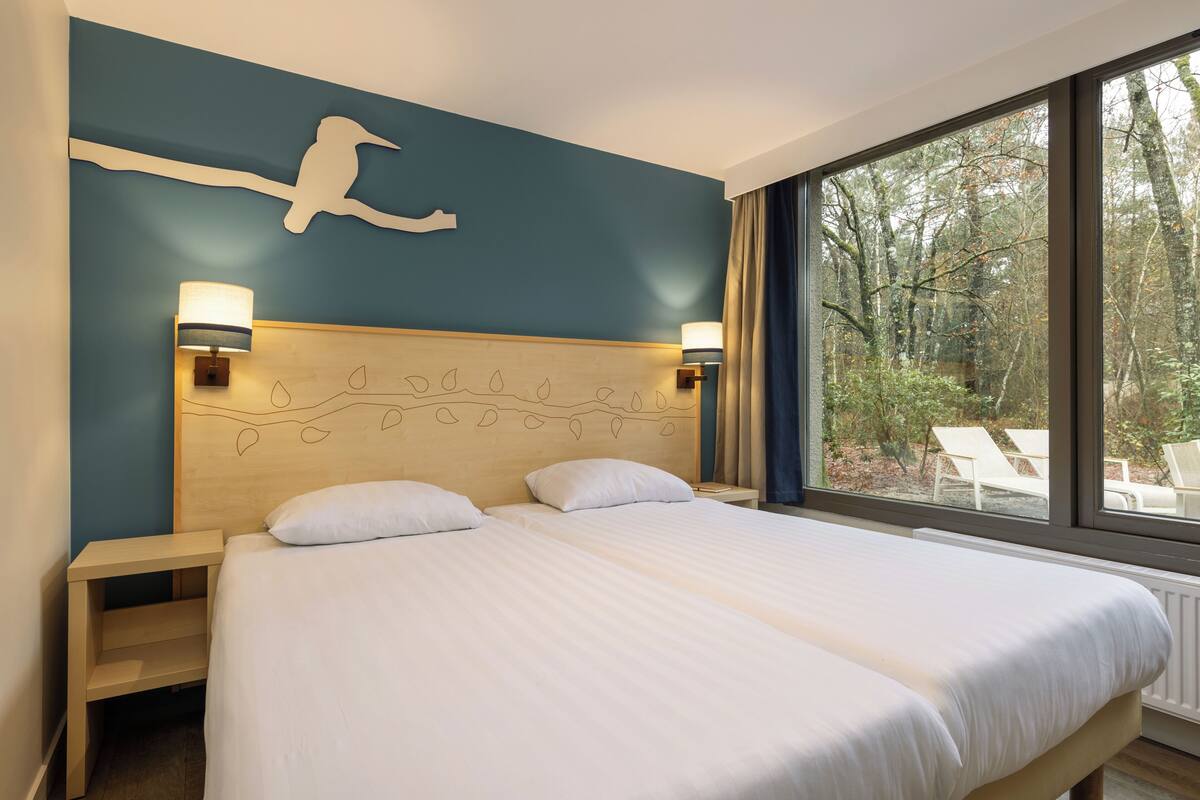 Una cama doble con sábanas blancas, cabecera de madera con diseños de hojas, dos lámparas de noche y vistas a un bosque a través de amplios ventanales.