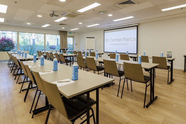 Una sala de conferencias con largas mesas, sillas y una pantalla de proyector que muestra "Center Parcs Meetings & Events".