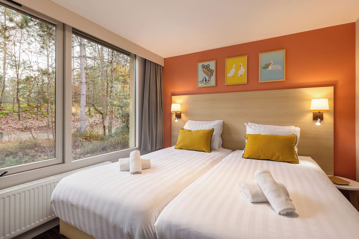 Habitación de hotel con dos camas, un ventanal con vista a árboles y lámparas fijadas a la pared.