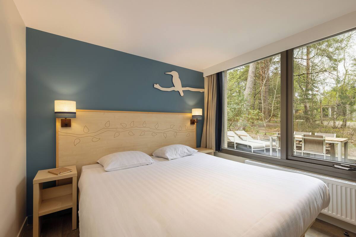Un dormitorio con una cama grande, dos mesitas de noche, una escultura de pájaro en la pared y vistas de un área de estar exterior a través de amplios ventanales.