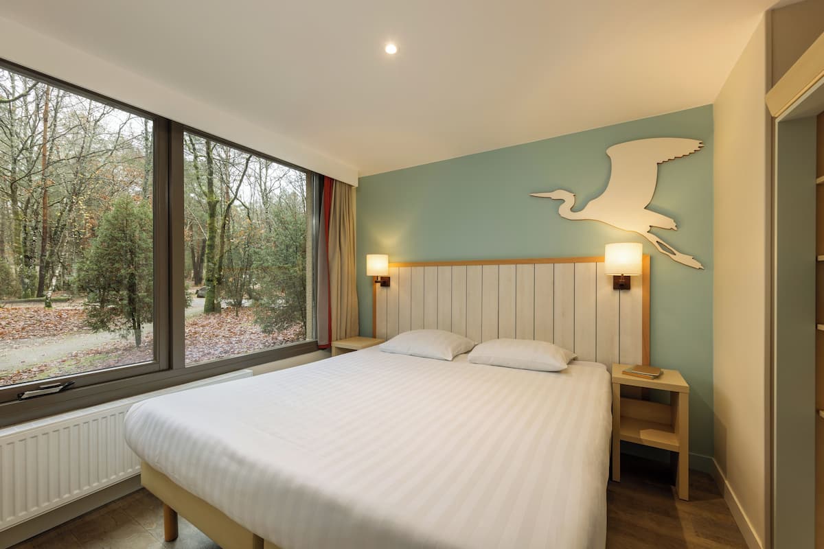Un dormitorio con un ventanal grande, una cama, una mesita de noche y una lámpara fijada a la pared.