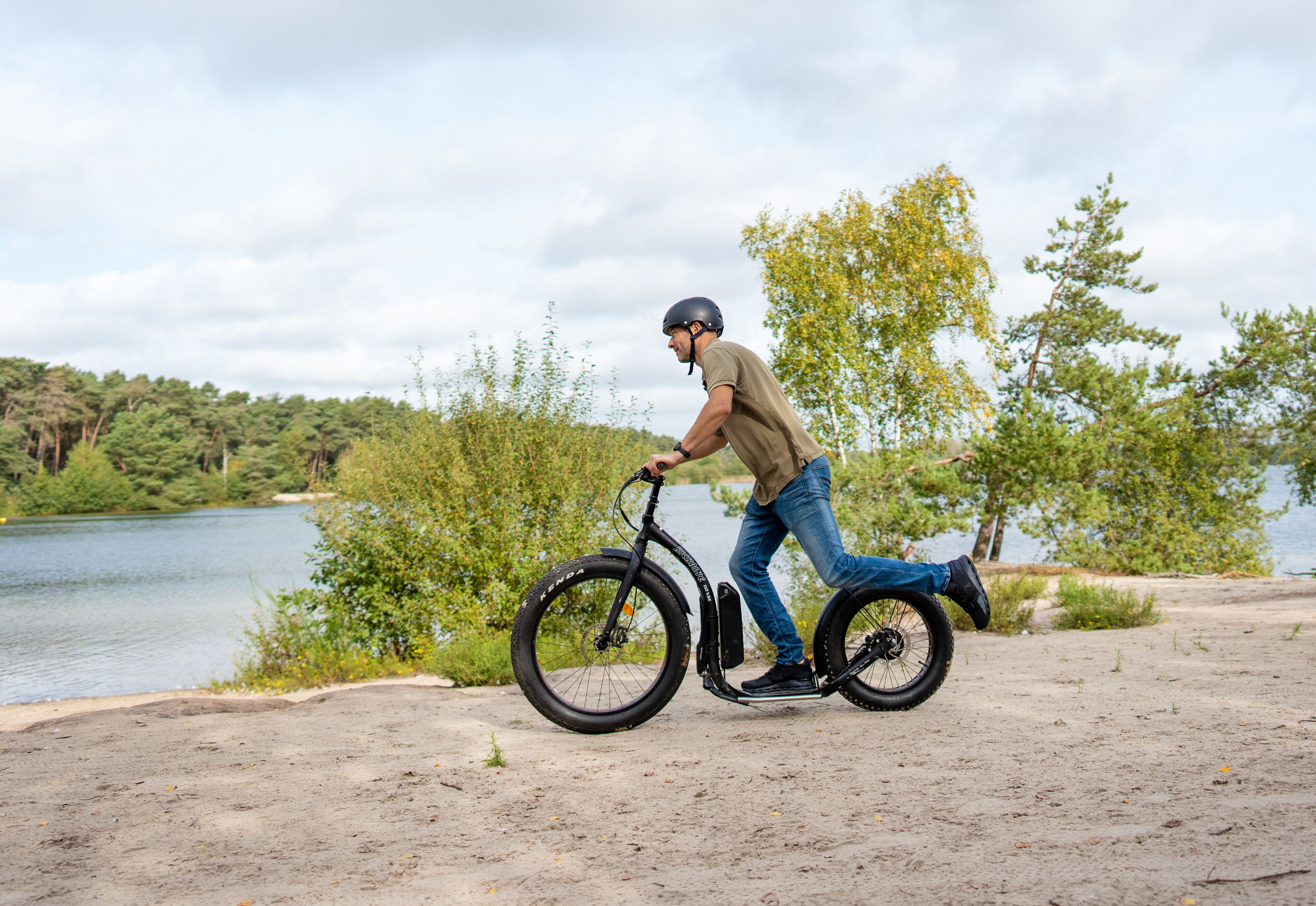 Een persoon fietst op een stilstaande fiets op een zandpad nabij een waterpartij met bomen op de achtergrond.