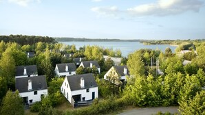 A cluster of white houses with dark roofs, surrounded by dense green trees, overlooking a calm lake. - Sunparks Kempense Meren (Mol)
