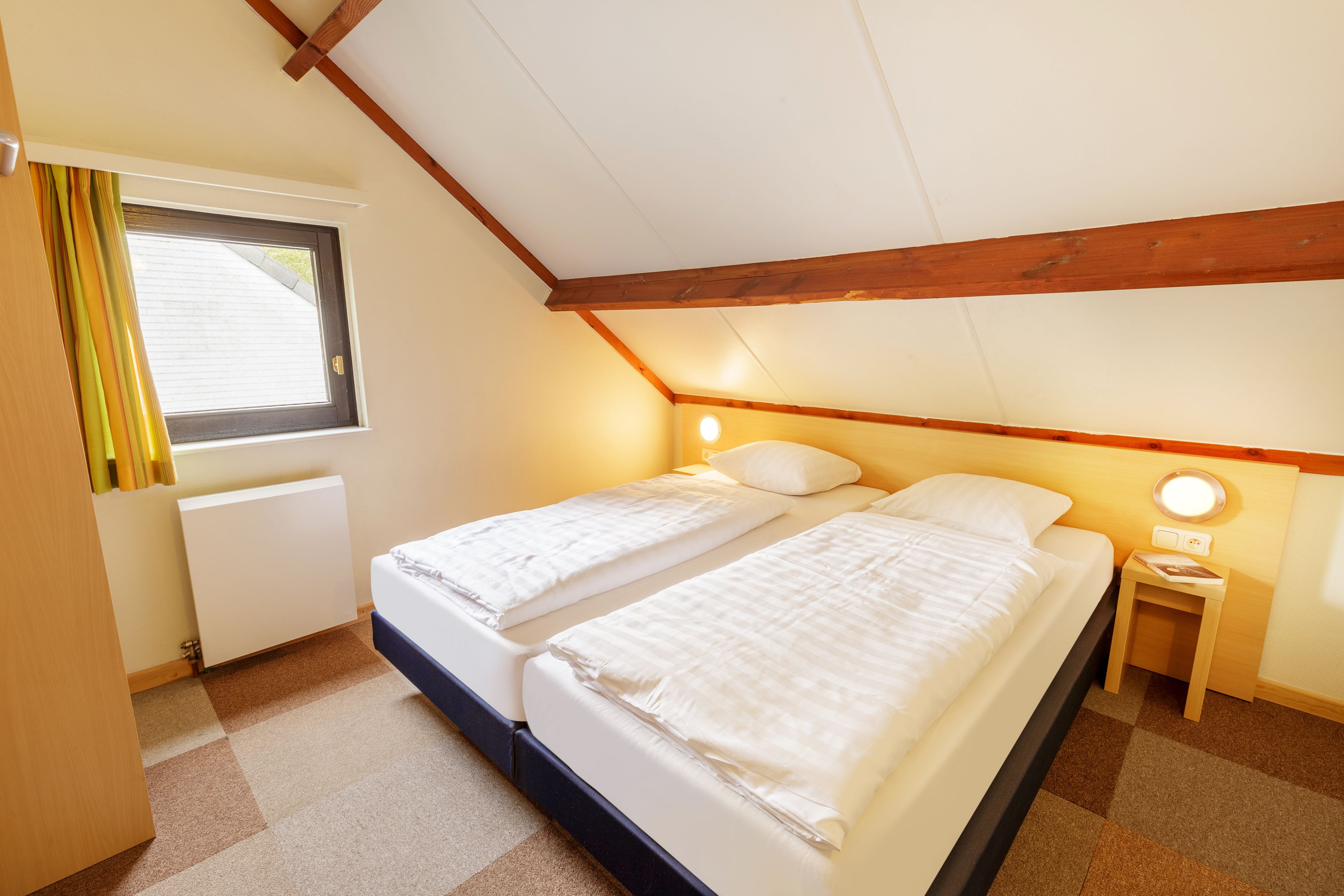 Een slaapkamer met twee bedden, een raam met gordijnen, een radiator en een nachtkastje met een lamp.