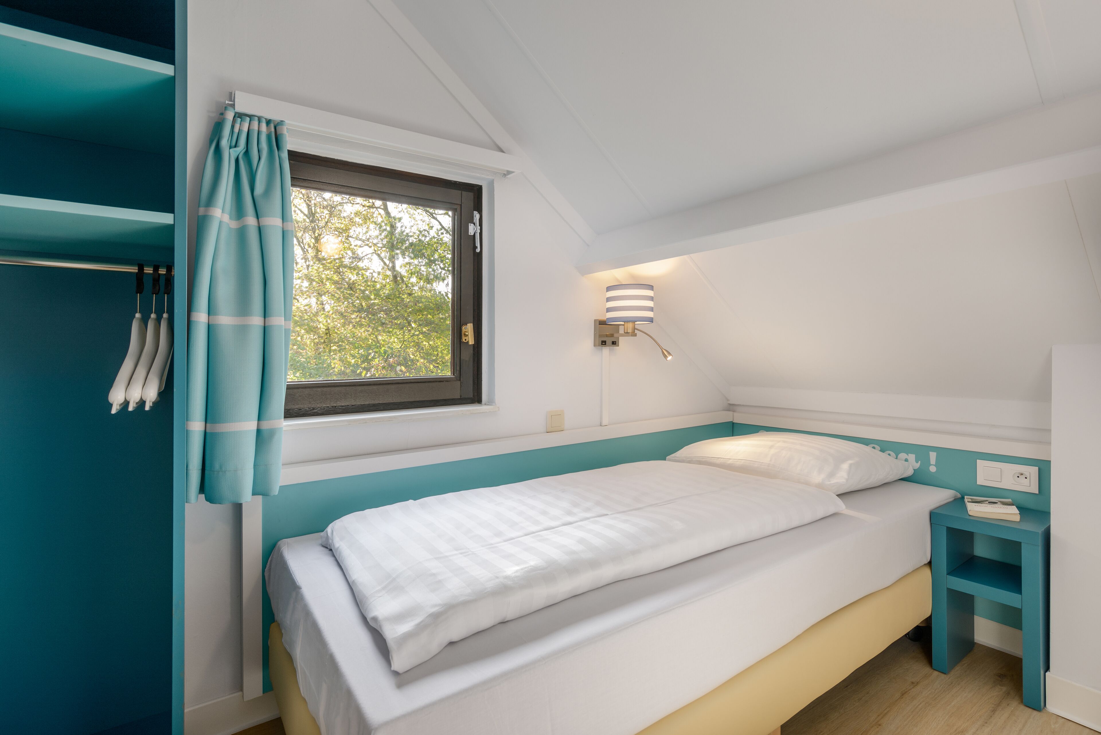 Een slaapkamer met een bed, een raam met gordijnen, een nachtkastje en een wandlamp.