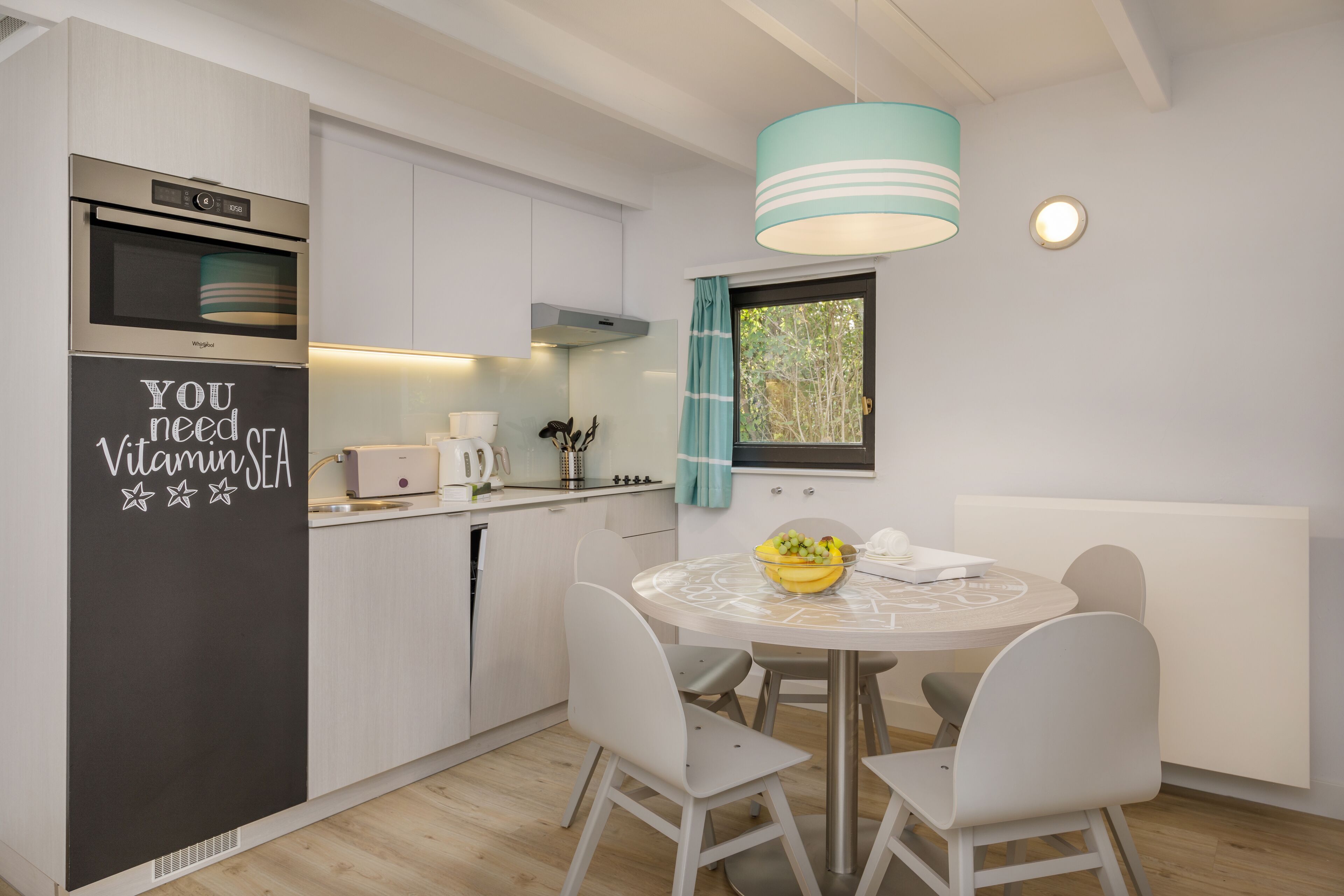 A modern kitchen with a built-in oven, a dining table with four chairs, and a window with curtains.
