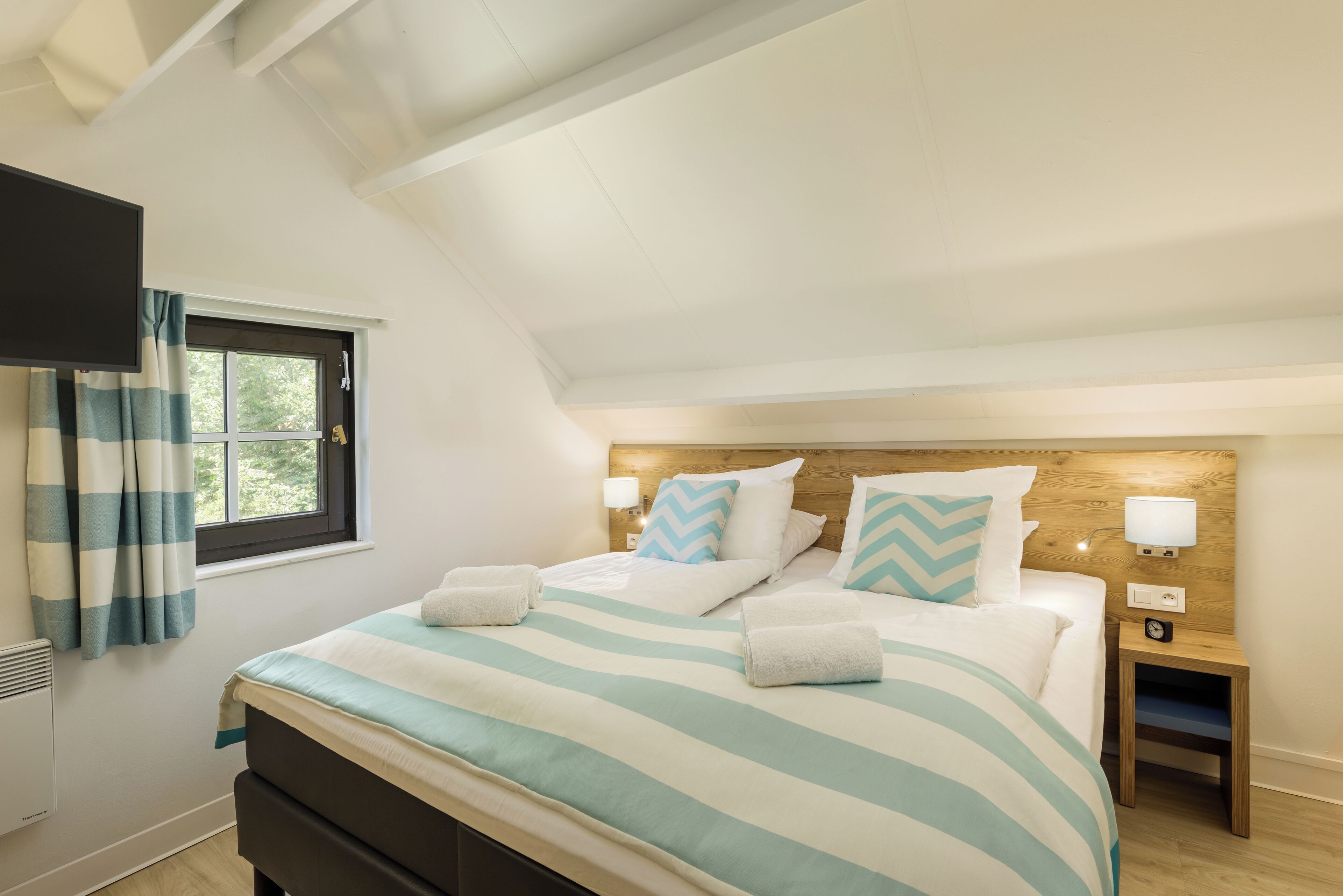Een slaapkamer met een bed, een nachtkastje, een aan de muur bevestigde televisie en een raam met gordijnen.