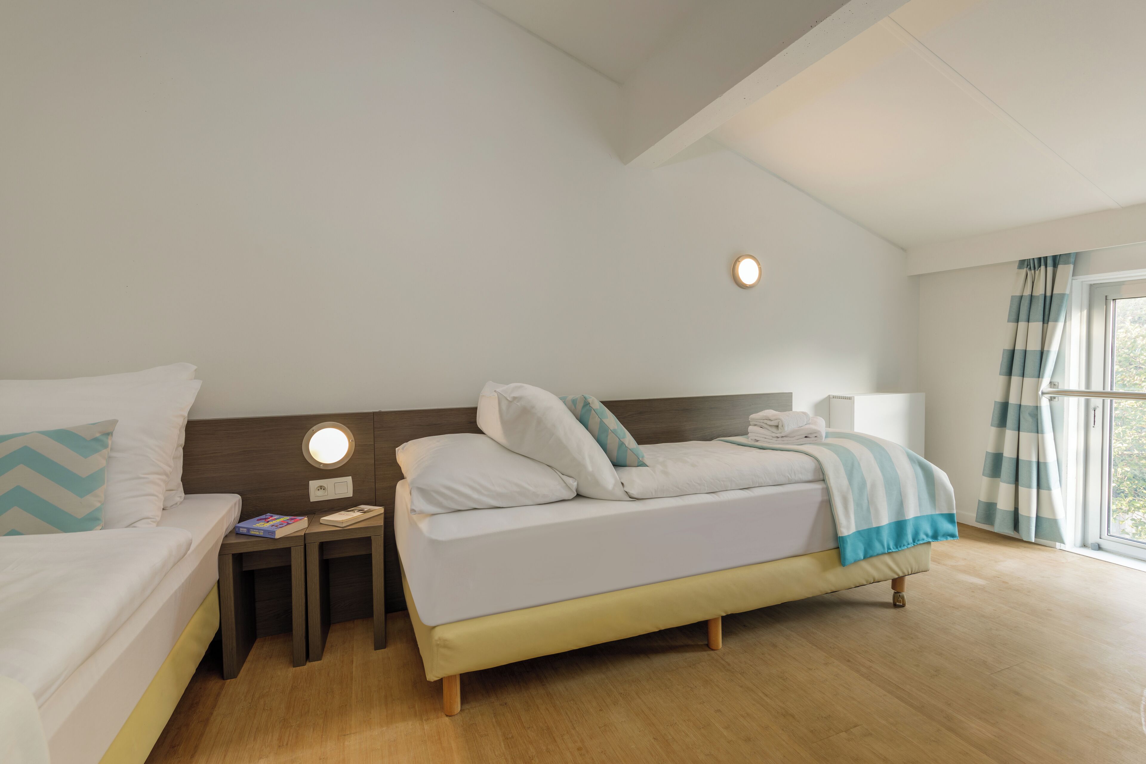 Ein Hotelzimmer mit zwei Betten, einem Nachttisch mit Lampe, einem Fenster mit Vorhängen und einer Deckenleuchte.