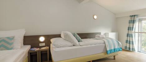 A hotel room with two beds, a bedside table with a lamp, a window with curtains, and a ceiling light.