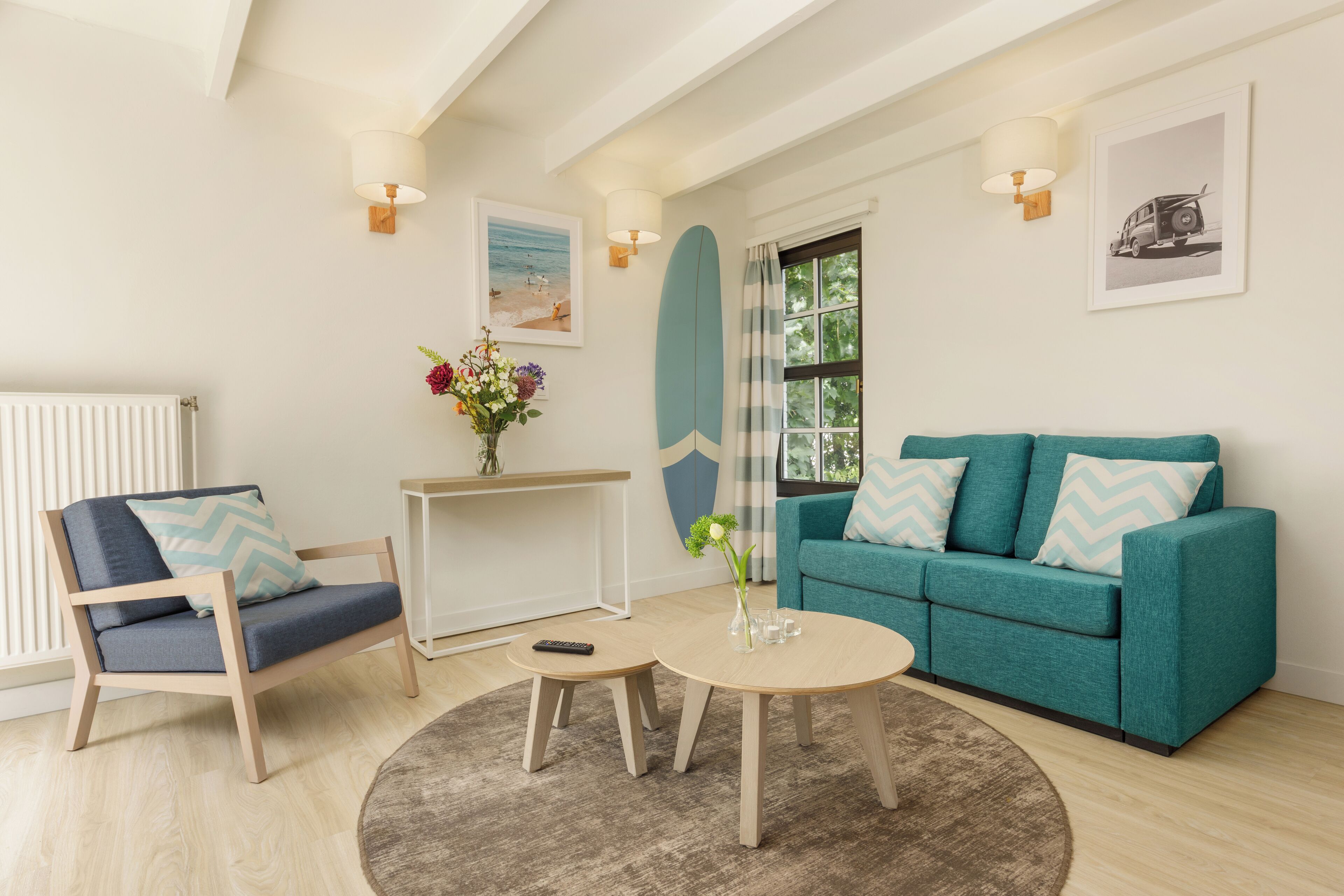 Een woonkamer met een blauwgroene bank, een houten stoel, een klein rond tafeltje en een salontafel. Er hangt een lijst met een afbeelding aan de muur en een surfboard decoratie.