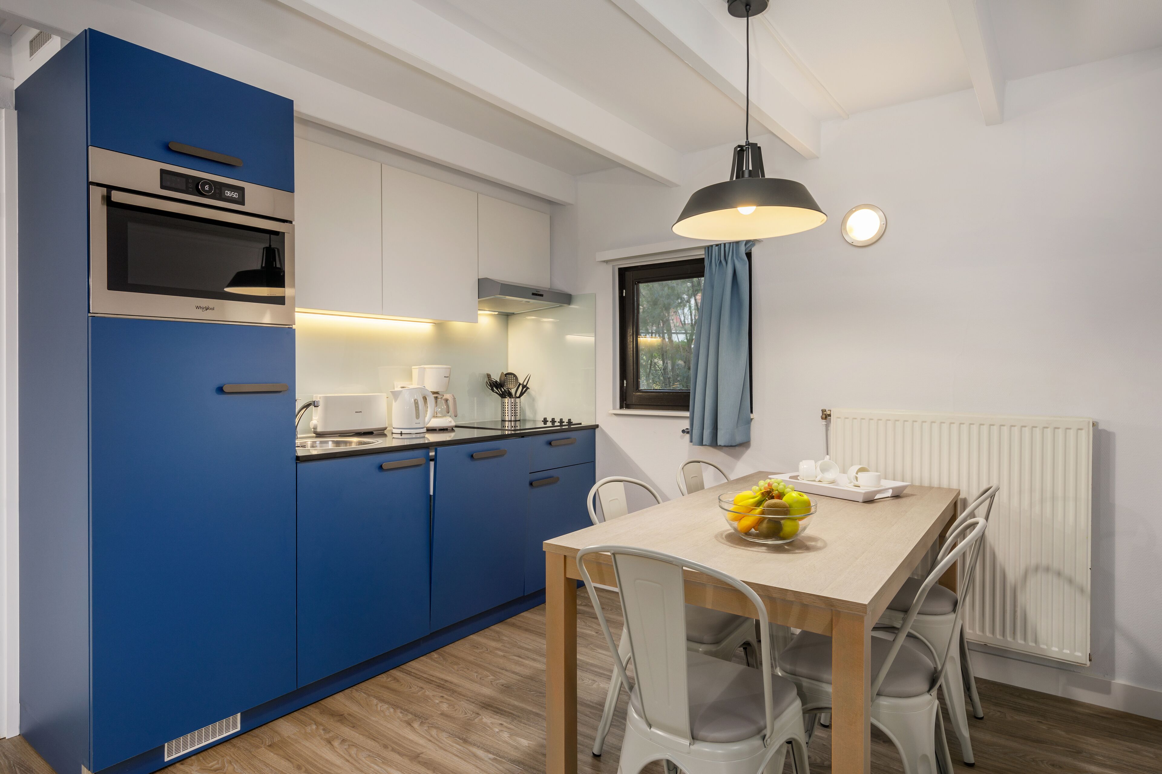 Une cuisine moderne avec des armoires bleues, un four encastré et un coin repas comprenant une table en bois et des chaises blanches.