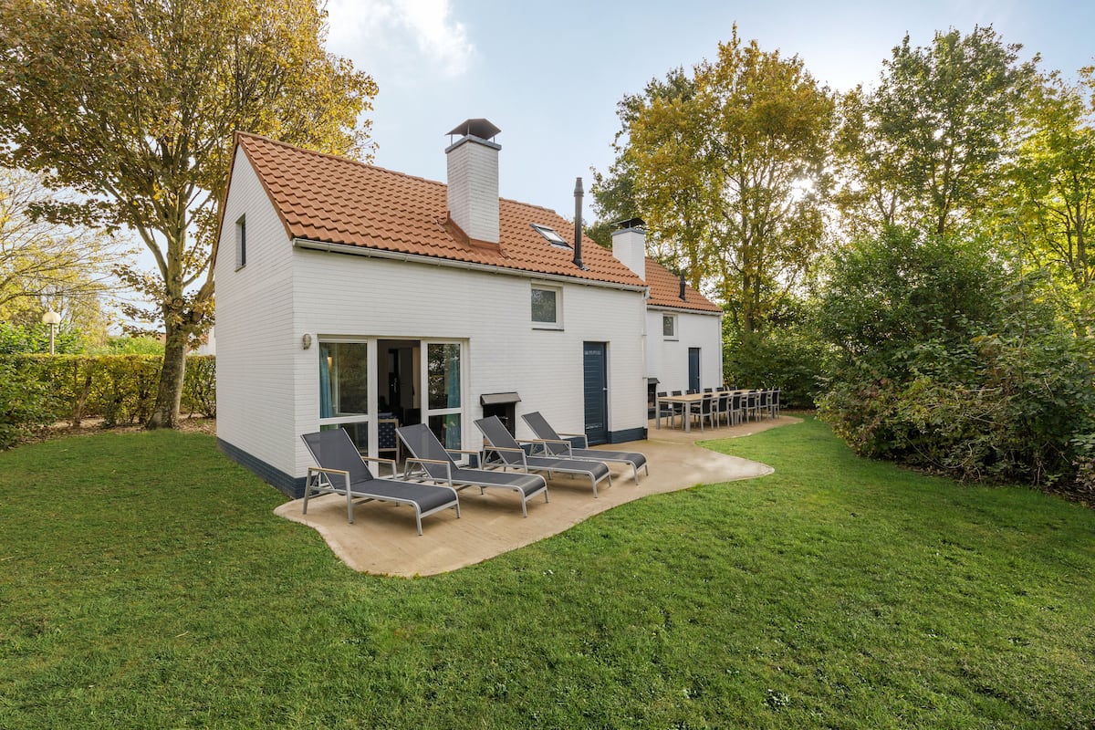 Une maison moderne avec un toit en tuiles, un patio couvert doté de chaises longues et une pelouse bien entretenue.