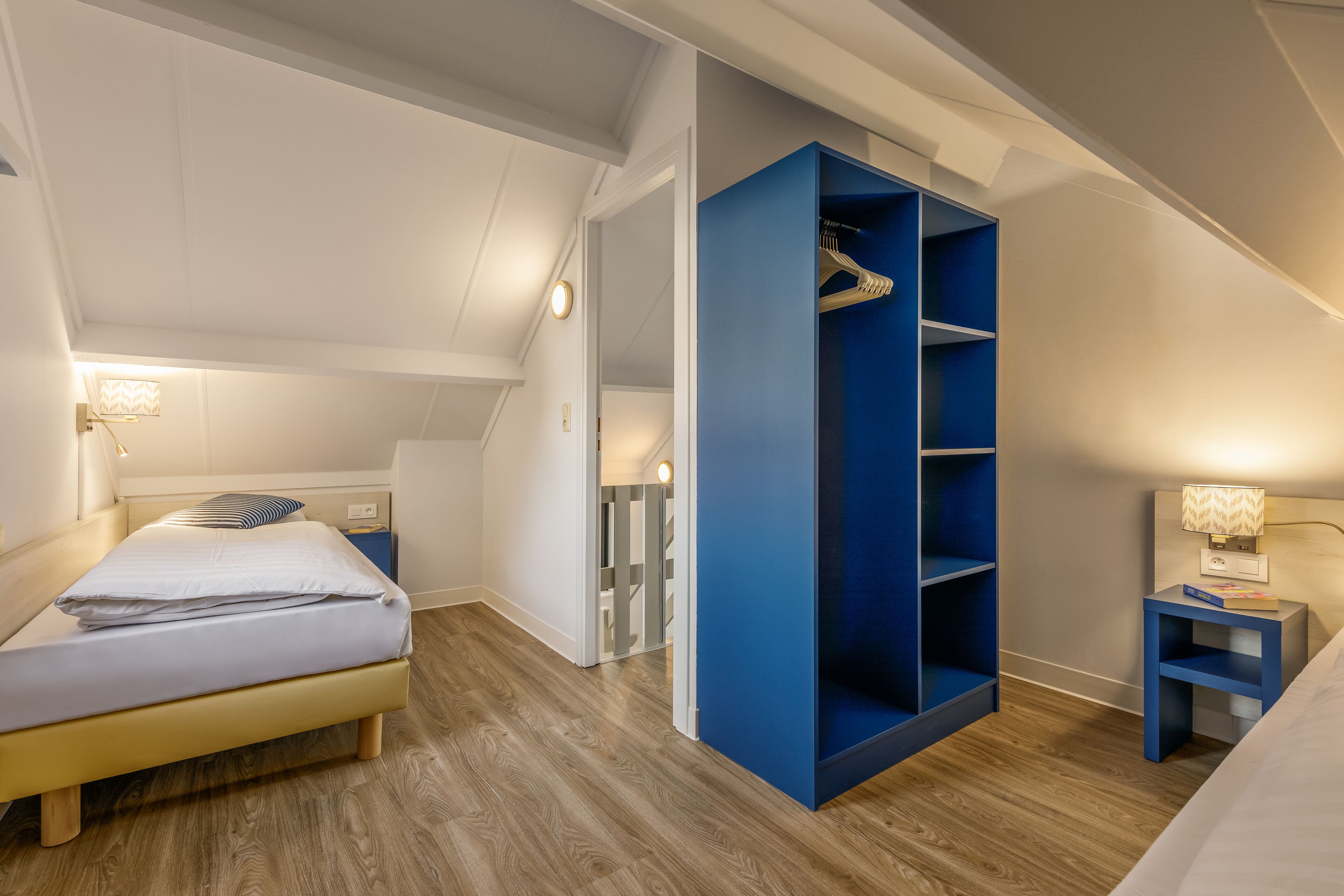 A bedroom with a bed, a blue wardrobe, a nightstand, and a staircase leading to another room.
