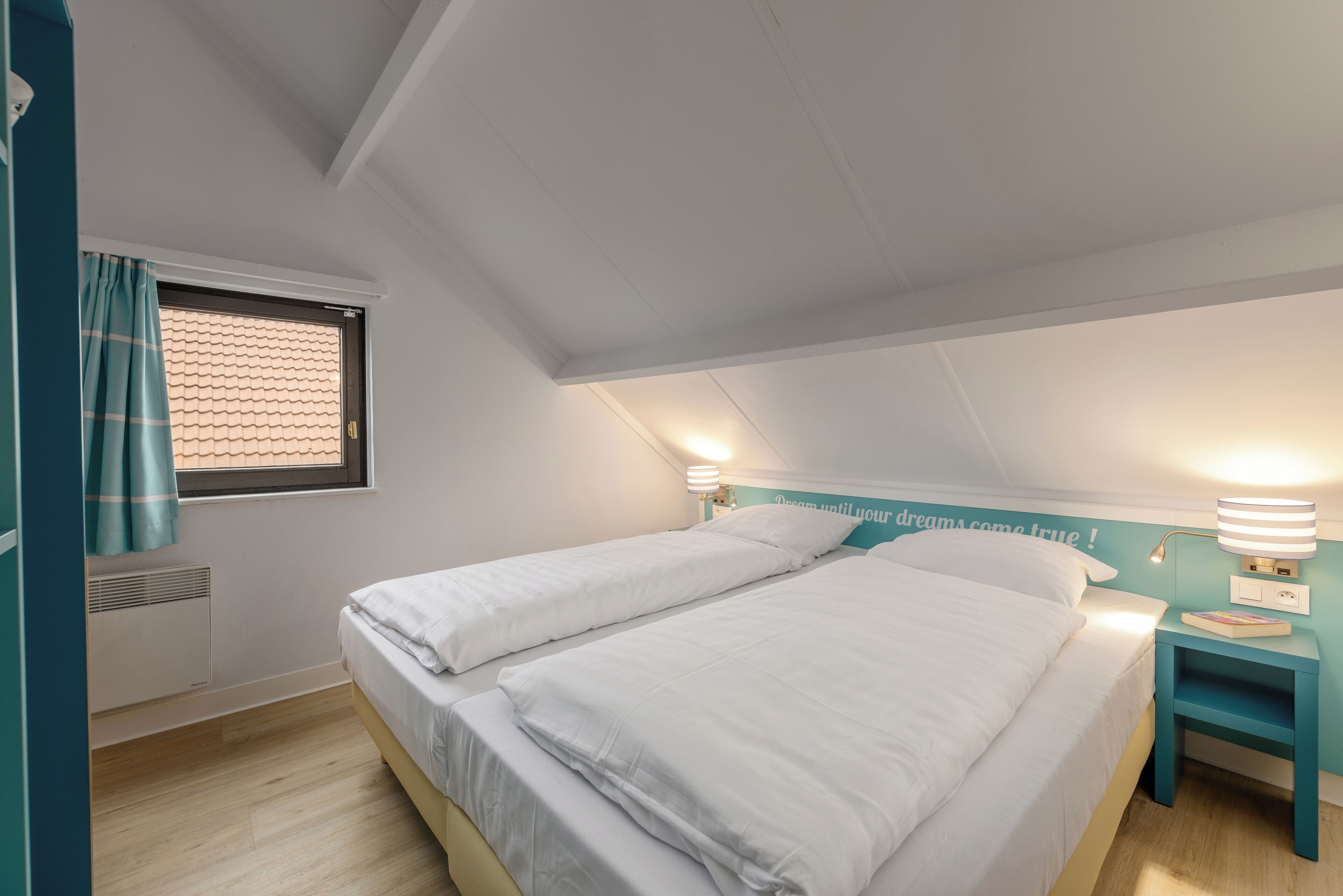 A bedroom with two beds, a window with blinds, a nightstand with a lamp, and a wall with a motivational quote.