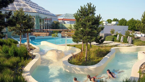 A pool area with a water slide, surrounded by greenery and a large building with a glass dome. - Center Parcs Park de Haan (De Haan)