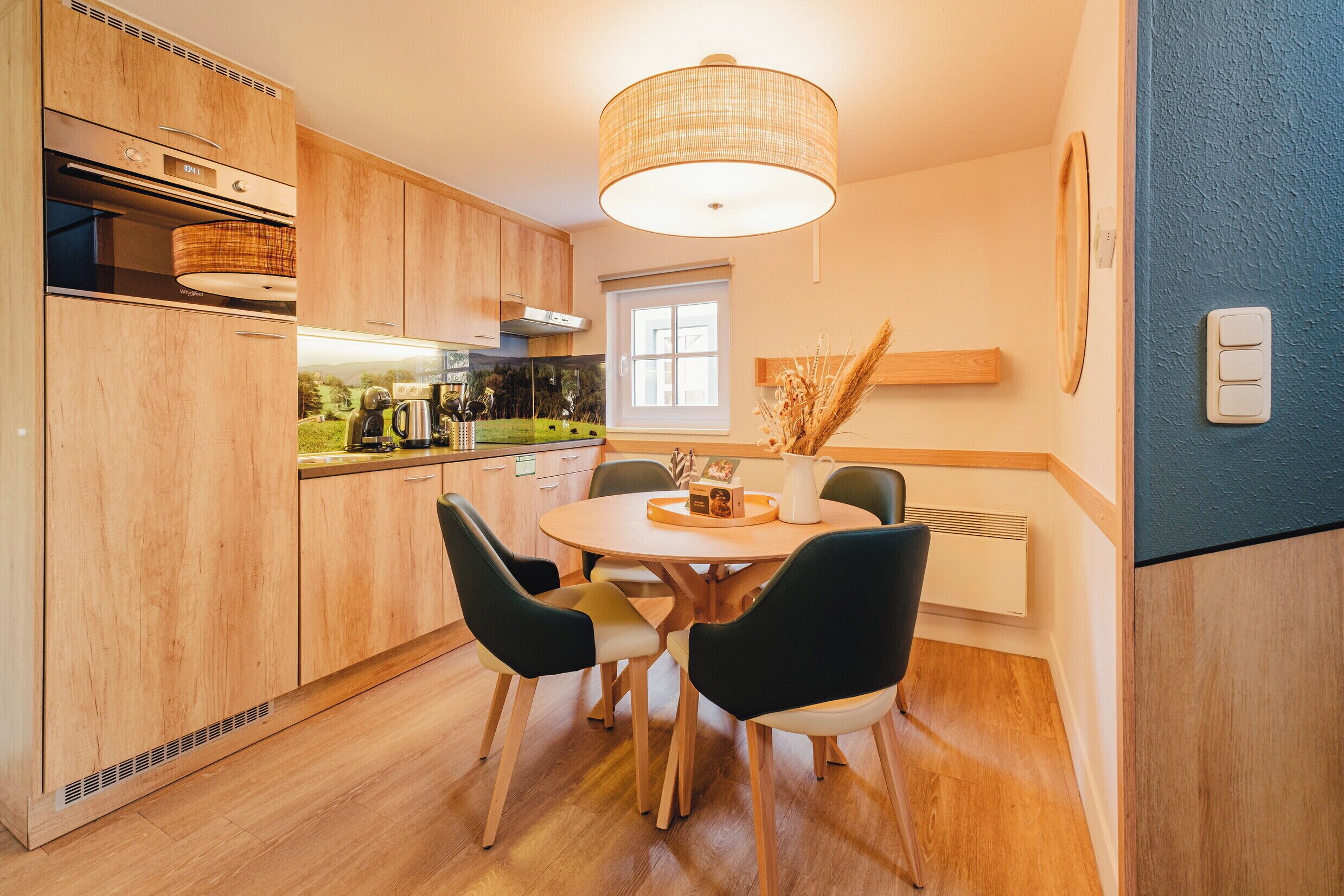 A modern kitchen with wooden cabinets, a round dining table with chairs, a hanging light fixture, and a window.