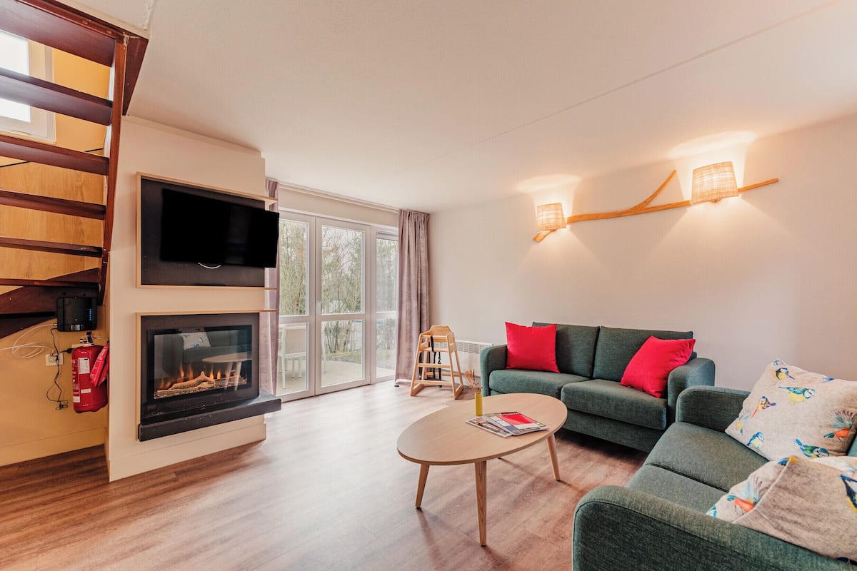 Un salon moderne avec une cheminée, un canapé vert agrémenté de coussins rouges, une table basse en bois et un escalier.