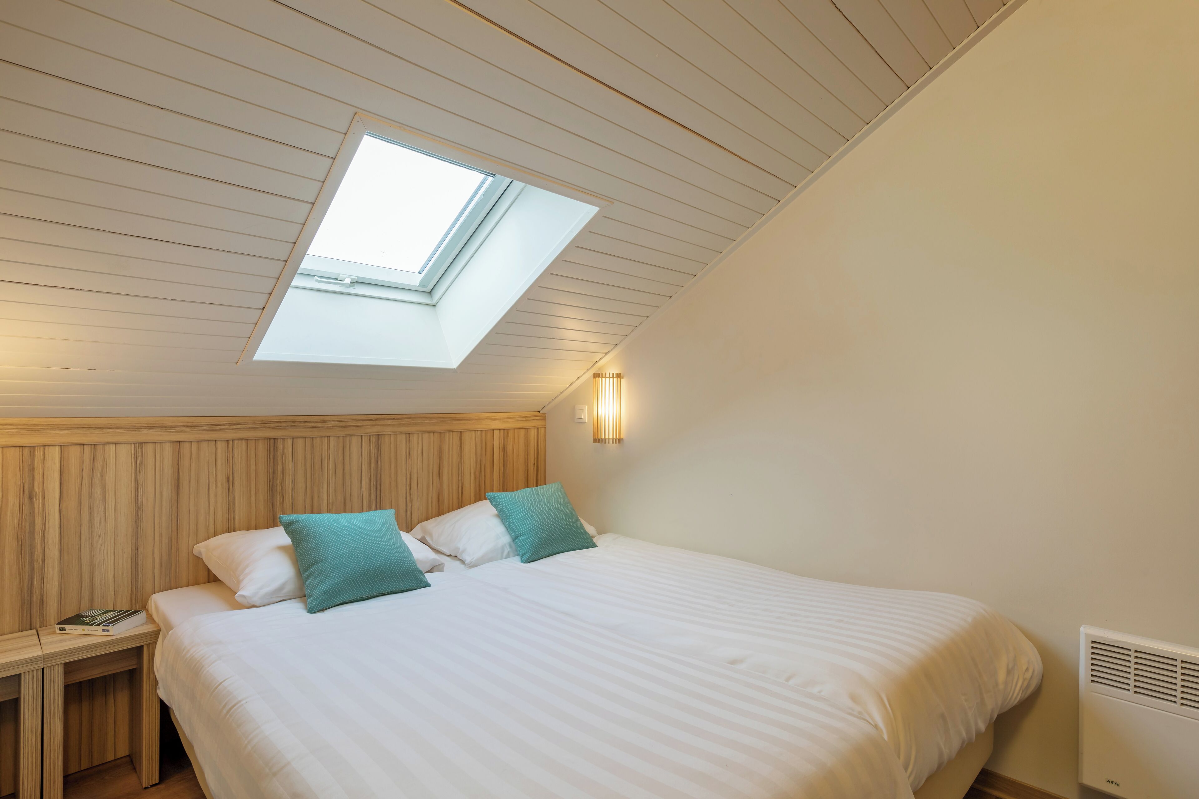 A bedroom with a skylight, a bed with white bedding and teal pillows, a bedside table with a book, and a wall-mounted light fixture.