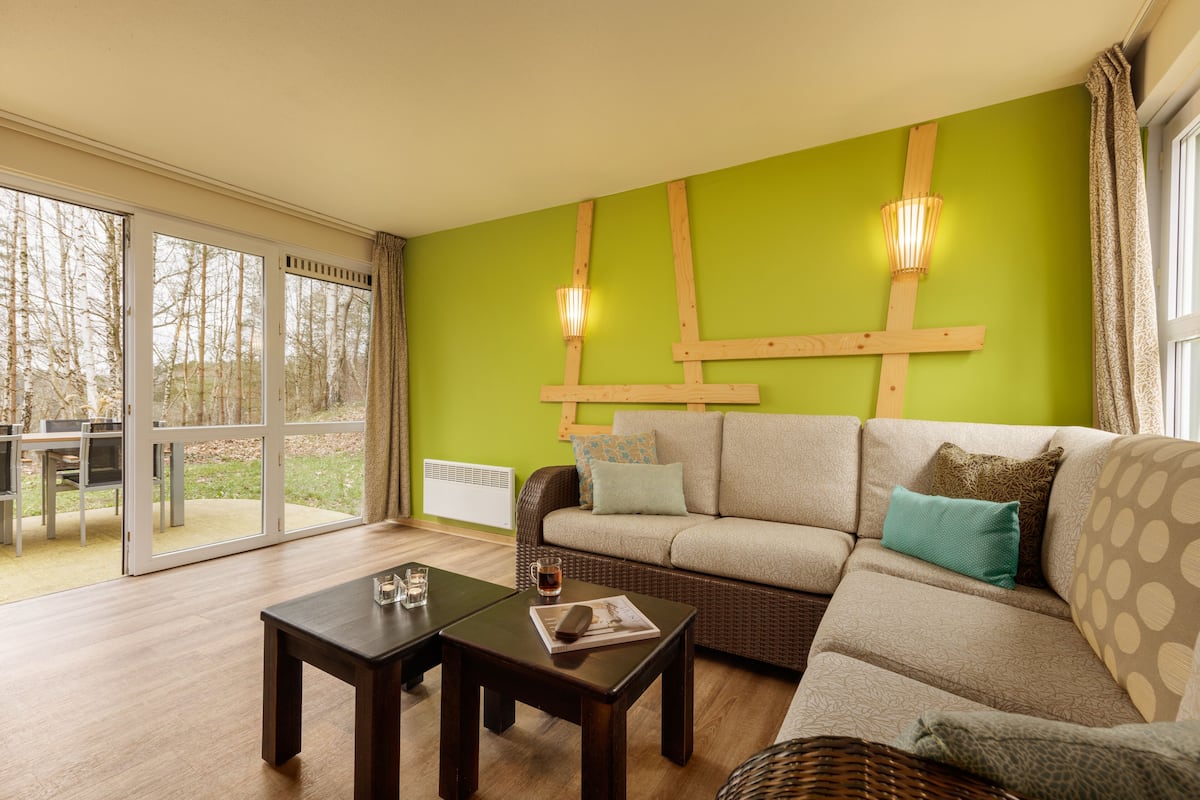 Un salon comprenant un canapé, deux tables basses, une cheminée et une grande fenêtre donnant sur la forêt.