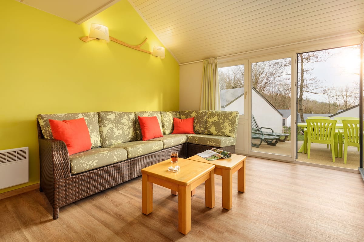 Une pièce lumineuse aux murs jaune vif, avec un canapé en osier, deux tables en bois et une porte-fenêtre coulissante donnant sur un espace extérieur agrémenté de chaises vertes.