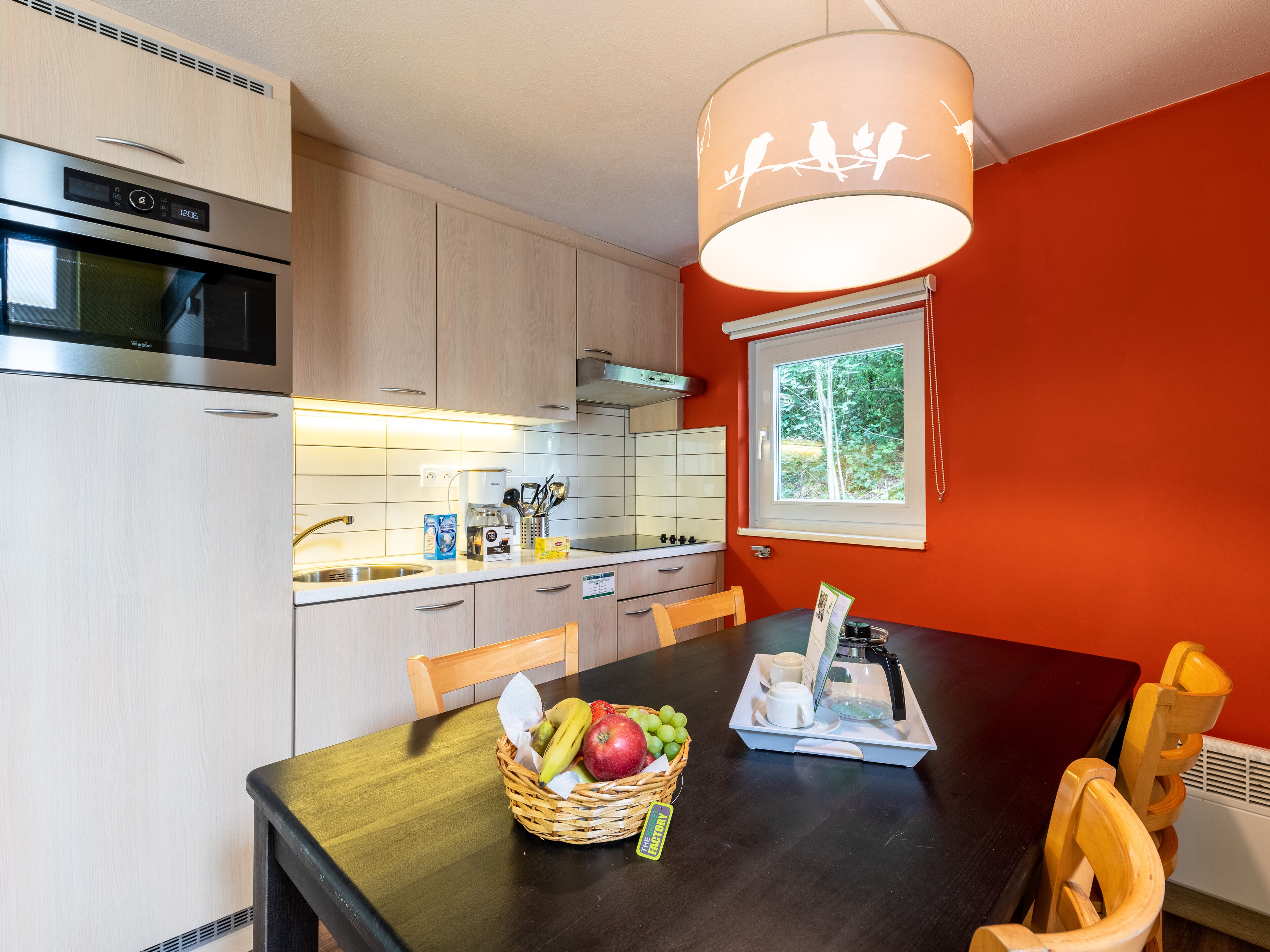 A modern kitchen with a dining area, featuring a refrigerator, oven, and a window with a view of trees.