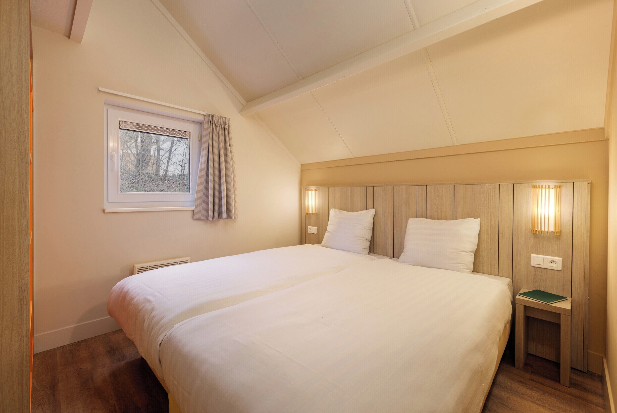 A bedroom with a large bed, a window with curtains, a bedside table with a lamp, and a wooden headboard.